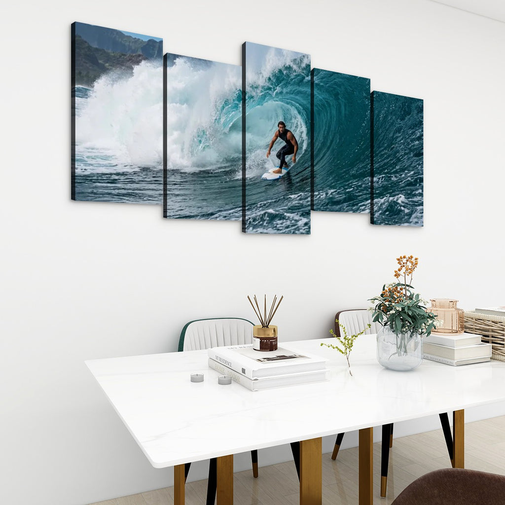 A five-panel canvas wall art set depicting a surfer riding a large blue ocean wave, displayed on a white wall above a modern white dining table with chairs and decorative items.