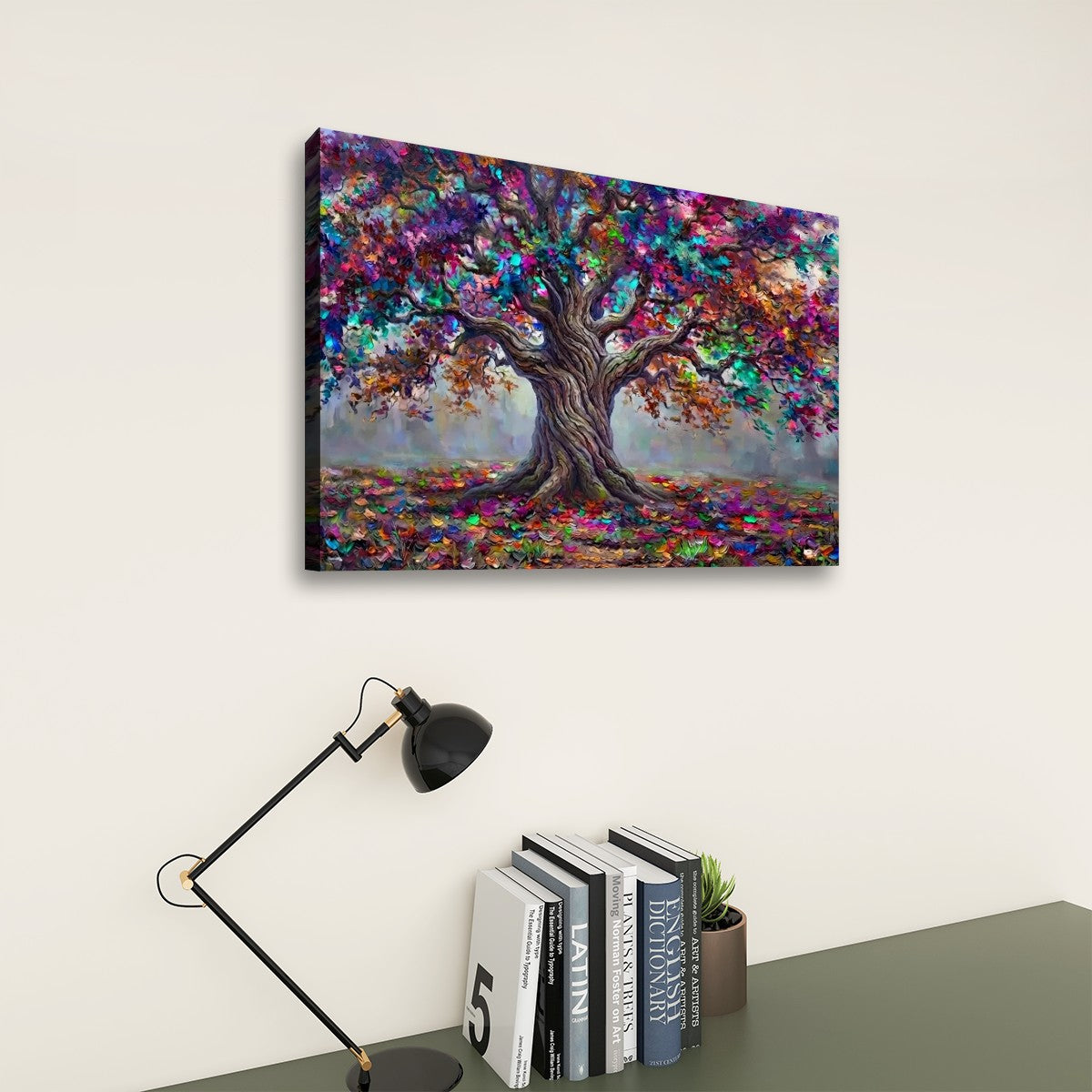 A colorful canvas wall art print depicting a stylized tree with vibrant multicolored leaves, shown hanging on a light beige wall above a desk with a black lamp and books.