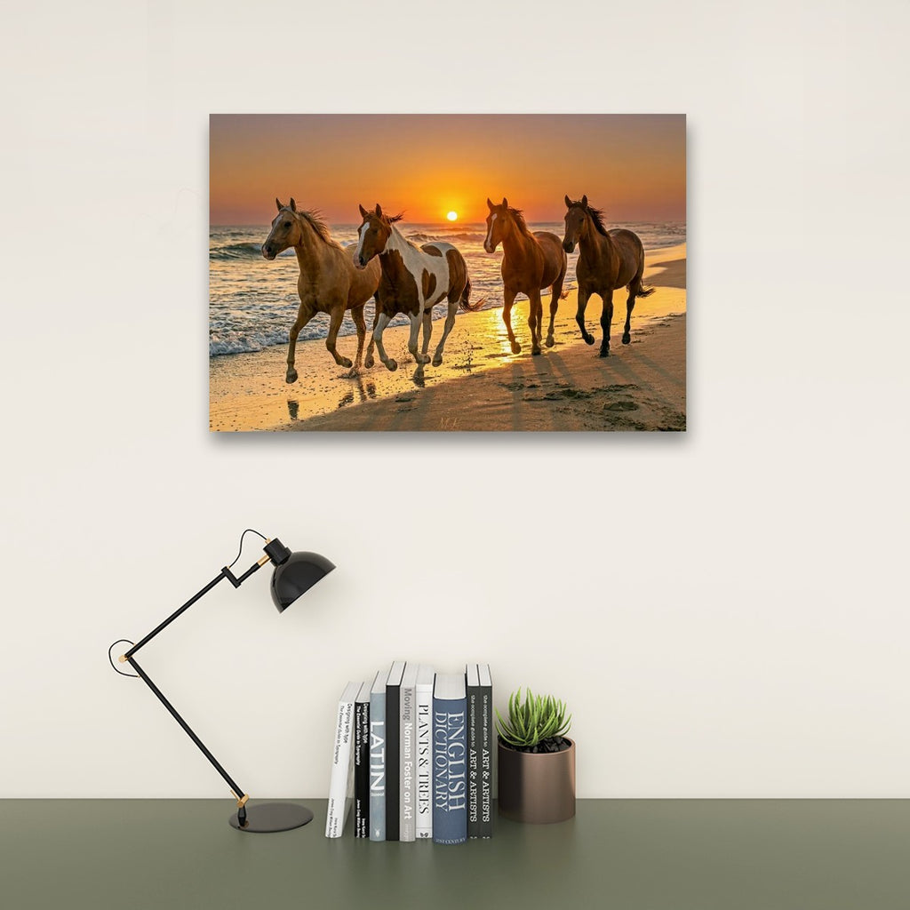 A rectangular wall art print showing four horses running on a beach at sunset, displayed above a desk with a black lamp, books, and a small potted succulent against a light wall.