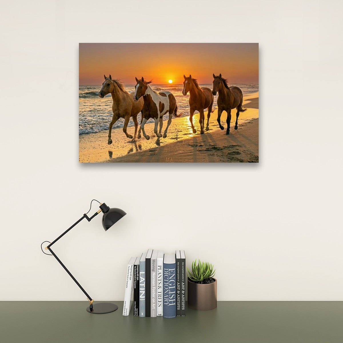 A rectangular wall art print showing four horses running on a beach at sunset, displayed above a desk with a black lamp, books, and a small potted succulent against a light wall.