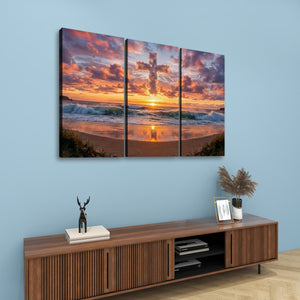 A three-panel canvas wall art set depicting a dramatic sunset over the ocean with a glowing cross in the sky, displayed above a wooden console table in a styled room with a light blue wall.