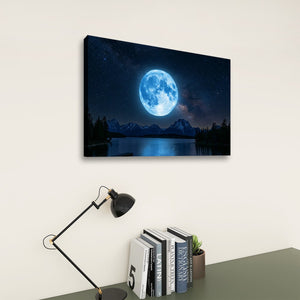 A rectangular canvas wall art print showing a large glowing blue moon over dark mountains and a reflective lake, displayed on a light wall above a desk with a black lamp and books.