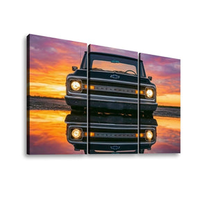 A three-panel canvas wall art print showing a black 1970 Chevy C10kfacing forward, reflected in water against a vivid orange and pink sunset sky. The panels are displayed as a triptych on a white background.