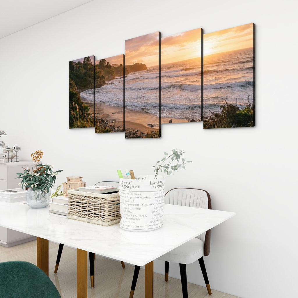 A 4-panel canvas wall art set depicting a beach sunset with waves, displayed on a white wall above a dining table in a modern room setting.