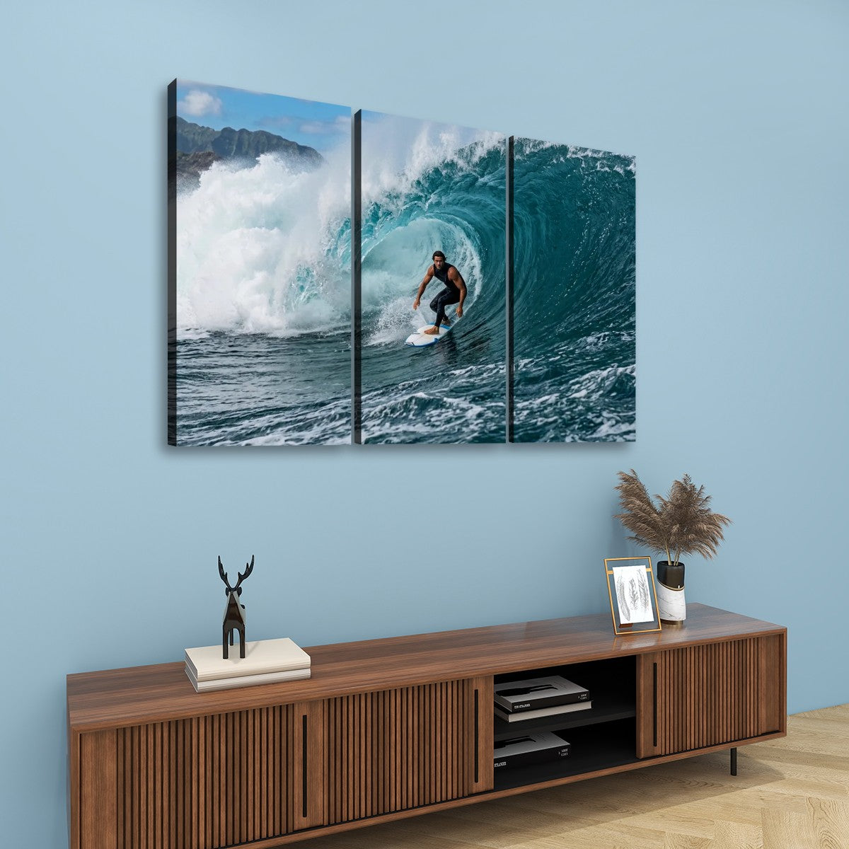 A three-panel canvas wall art set depicting a surfer riding a large blue ocean wave, displayed on a light blue wall above a wooden console table with decorative items.