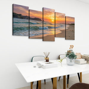 A five-panel canvas wall art set depicting a beach sunset with ocean waves, displayed above a white dining table in a modern room setting.