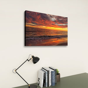 A rectangular wall art print showing a dramatic orange and red sunset over ocean waves, displayed above a desk with a black lamp and books against a light wall.