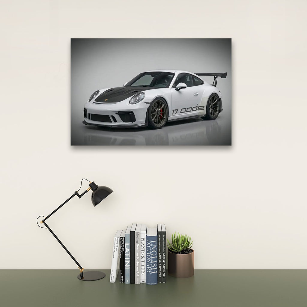 A framed wall art print showing a white Porsche 911 Carrera RSR with a black hood and rear wing, displayed above a desk with a black lamp, books, and a small potted succulent against a light beige wall.