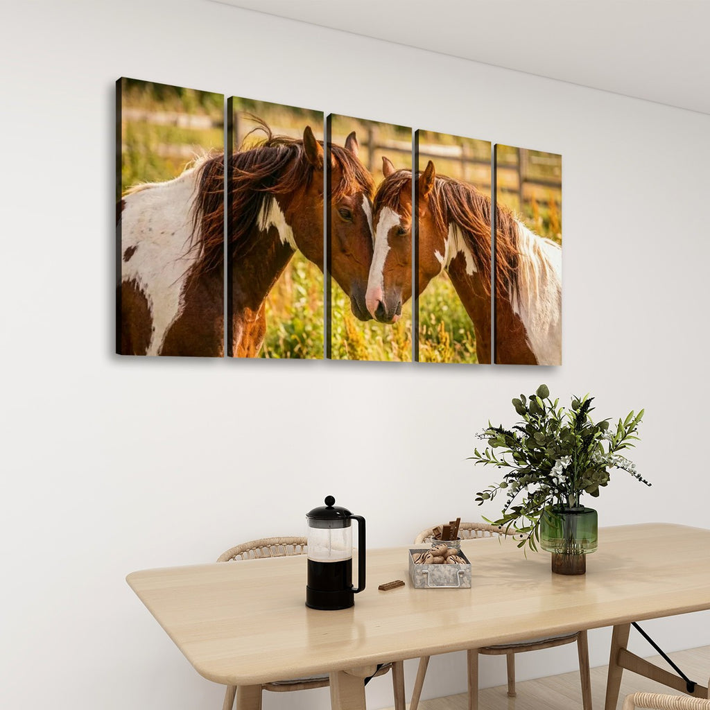 A five-panel canvas wall art set depicting two brown and white horses in a sunlit field, displayed on a white wall above a light wood dining table with a vase of greenery and a French press.