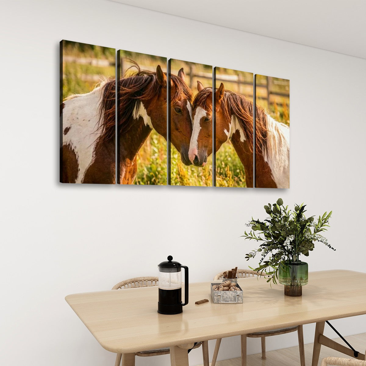 A five-panel canvas wall art set depicting two brown and white horses in a sunlit field, displayed on a white wall above a light wood dining table with a vase of greenery and a French press.