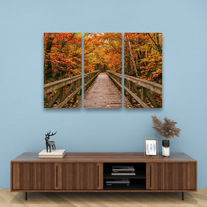 A three-panel canvas wall art set depicting a wooden boardwalk bridge over a vibrant autumn forest with orange and red foliage, displayed on a light blue wall above a modern brown wooden media console.
