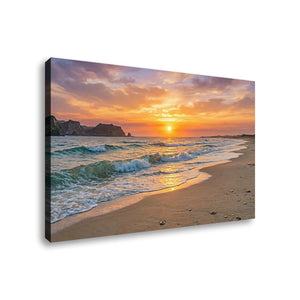A rectangular canvas wall art print showing a beach sunset scene with ocean waves, sandy shore, cliffs, and a glowing sun in the sky, photographed against a plain white background.