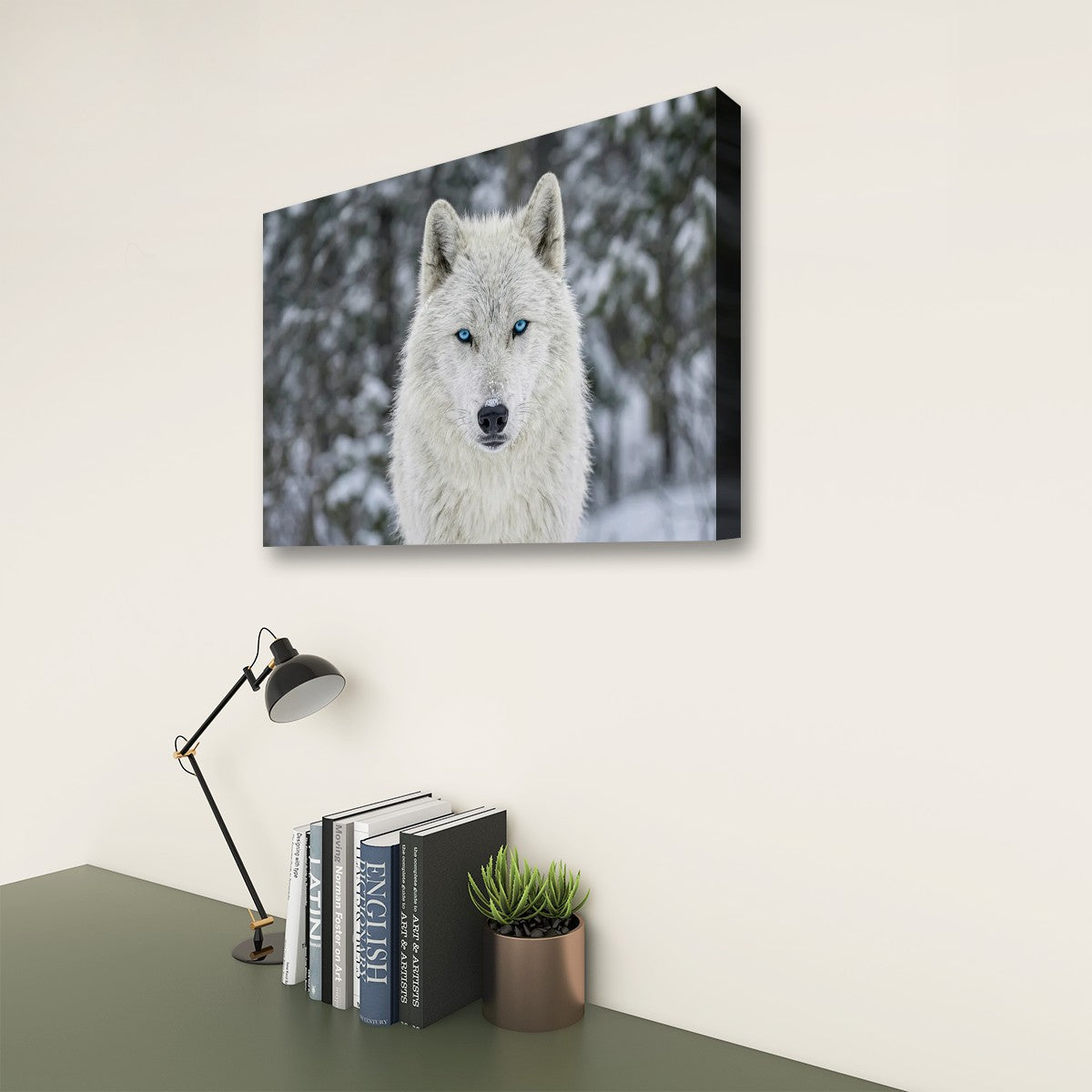A rectangular wall art print showing a close-up portrait of a white wolf with blue eyes in a snowy outdoor setting, displayed on a beige wall above a desk with books, a lamp, and a small plant.
