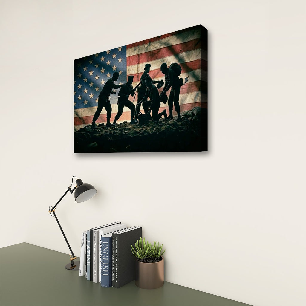 A rectangular wall art print showing silhouetted soldiers carrying a flag across rocky ground, displayed above a desk with a lamp, books, and a small plant in a real interior setting.