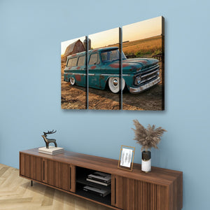 A three-panel canvas wall art print depicting a vintage teal 1966 Chevy Suburban Carryall in a rural field at sunset, displayed on a light blue wall above a wooden console table with decorative items.