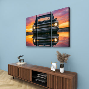A three-panel canvas wall art print depicting a black vintage 1970 Chevy C10 reflected in water at sunset, displayed on a light blue wall above a wooden console table with decorative items.