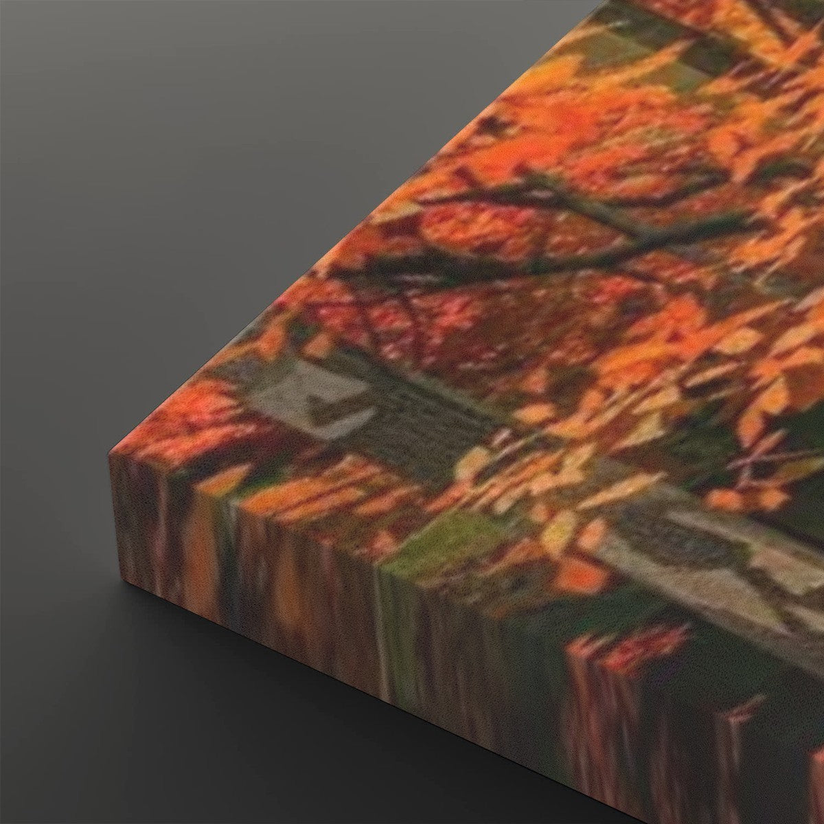 A close-up of a rectangular canvas or print showing an autumn forest scene with orange and red leaves, dark branches, and a blurred reflective Huay Mae Khamin Falls, photographed on a dark gray background.