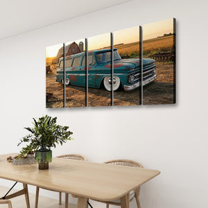 A five-panel canvas wall art print depicting a vintage teal 1966 Chevy Suburban Carryall station wagon parked in a rural field at sunset, displayed on a white wall above a wooden dining table with chairs and a plant.