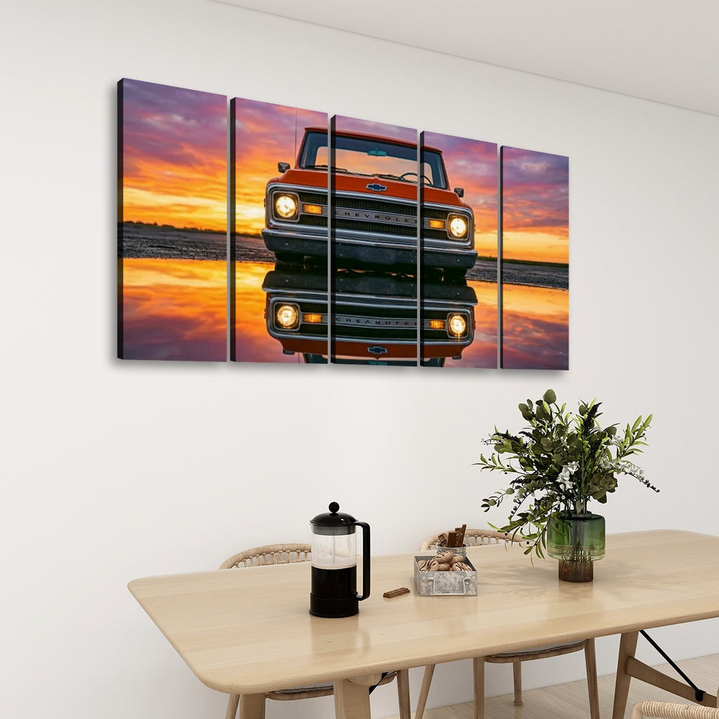 A 4-panel canvas wall art set depicting a red vintage pickup truck reflected in water at sunset, displayed on a white wall above a dining table with a vase and coffee maker.