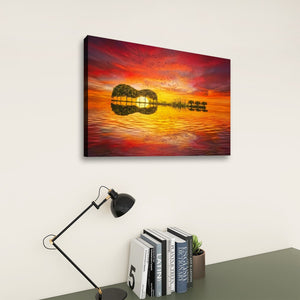 A rectangular wall art print showing a vivid sunset over water with tree silhouettes, displayed above a desk with a black lamp and books against a light wall.
