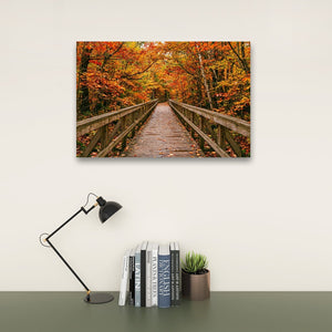 A rectangular wall art print showing a wooden boardwalk bridge over water surrounded by autumn trees with orange and yellow leaves, displayed above a desk with a black lamp, books, and a small potted succulent against a light wall.