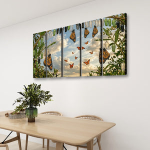 A four-panel canvas wall art set depicting monarch butterflies hanging from a tree with green leaves and a cloudy sky, displayed on a white wall above a light wood dining table with chairs and a potted plant.