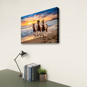 A rectangular wall art print showing three horses running along a beach at sunset, displayed on a light wall above a desk with a lamp, books, and a small plant.