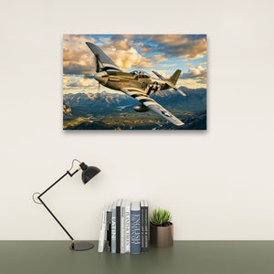 A rectangular wall art print showing a vintage military airplane flying over mountains under a cloudy sky, displayed above a desk with a black lamp, books, and a small potted succulent against a light wall.