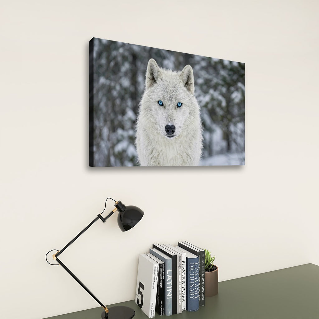 A rectangular wall art print showing a white wolf with bright blue eyes in a snowy outdoor setting, displayed above a desk with books, a small plant, and a black desk lamp against a light beige wall.