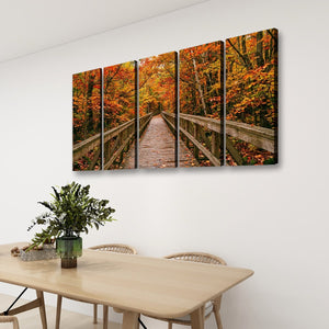 A four-panel canvas wall art set depicting an autumn forest bridge with orange and red foliage, displayed on a white wall above a dining table with chairs and a plant.