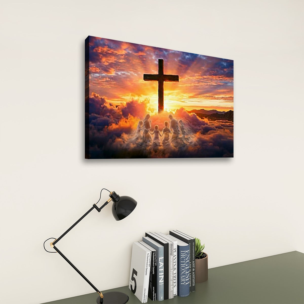 A rectangular wall art print showing a dark cross silhouette against a dramatic sunset sky with clouds and glowing light, displayed on a light wall above a desk with a lamp, books, and a small plant.