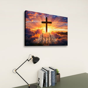 A rectangular wall art print showing a dark cross silhouette against a dramatic sunset sky with clouds and glowing light, displayed on a light wall above a desk with a lamp, books, and a small plant.