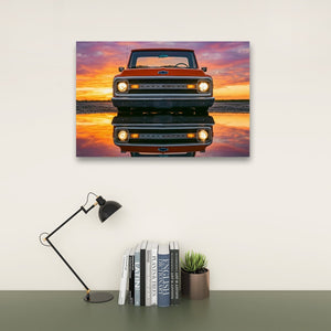 A rectangular wall art print showing the front of an orange  1970 Chevrolet C10 truck reflected in water at sunset, displayed above a desk with a black lamp, books, and a small succulent in a styled interior scene.