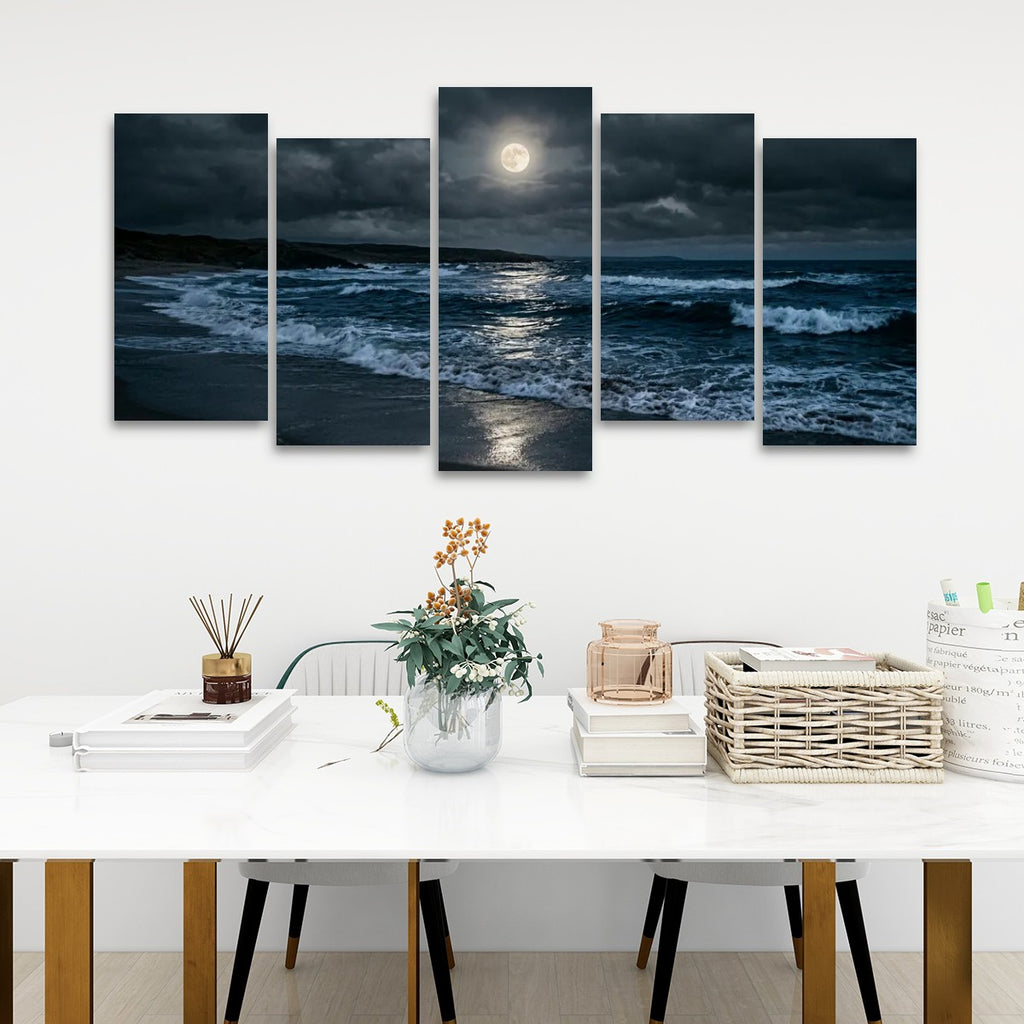 A five-panel canvas wall art set depicting a dramatic ocean scene at night under a full moon, displayed on a white wall above a styled dining table with books, a vase, and a basket.