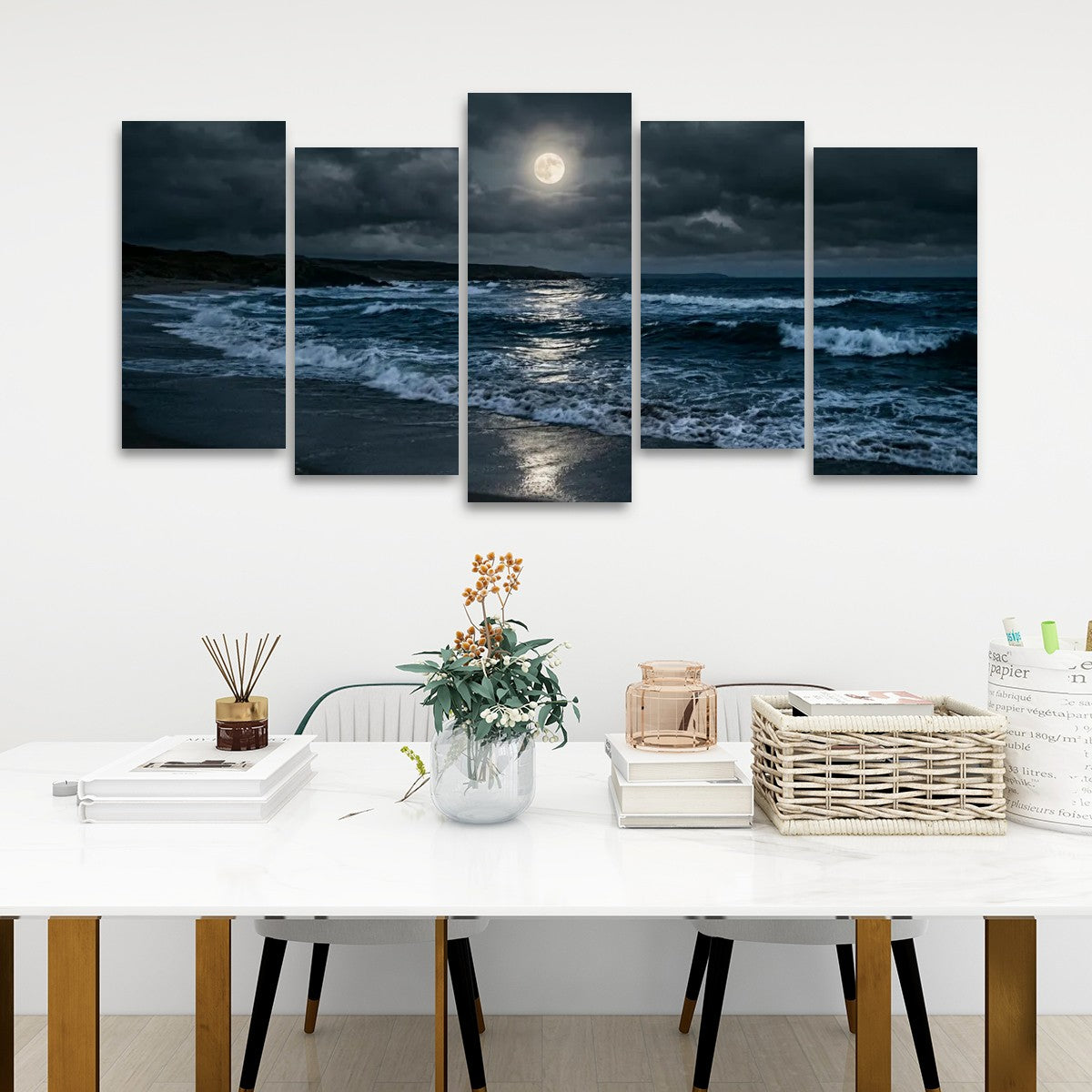 A five-panel canvas wall art set depicting a dramatic ocean scene at night under a full moon, displayed on a white wall above a styled dining table with books, a vase, and a basket.