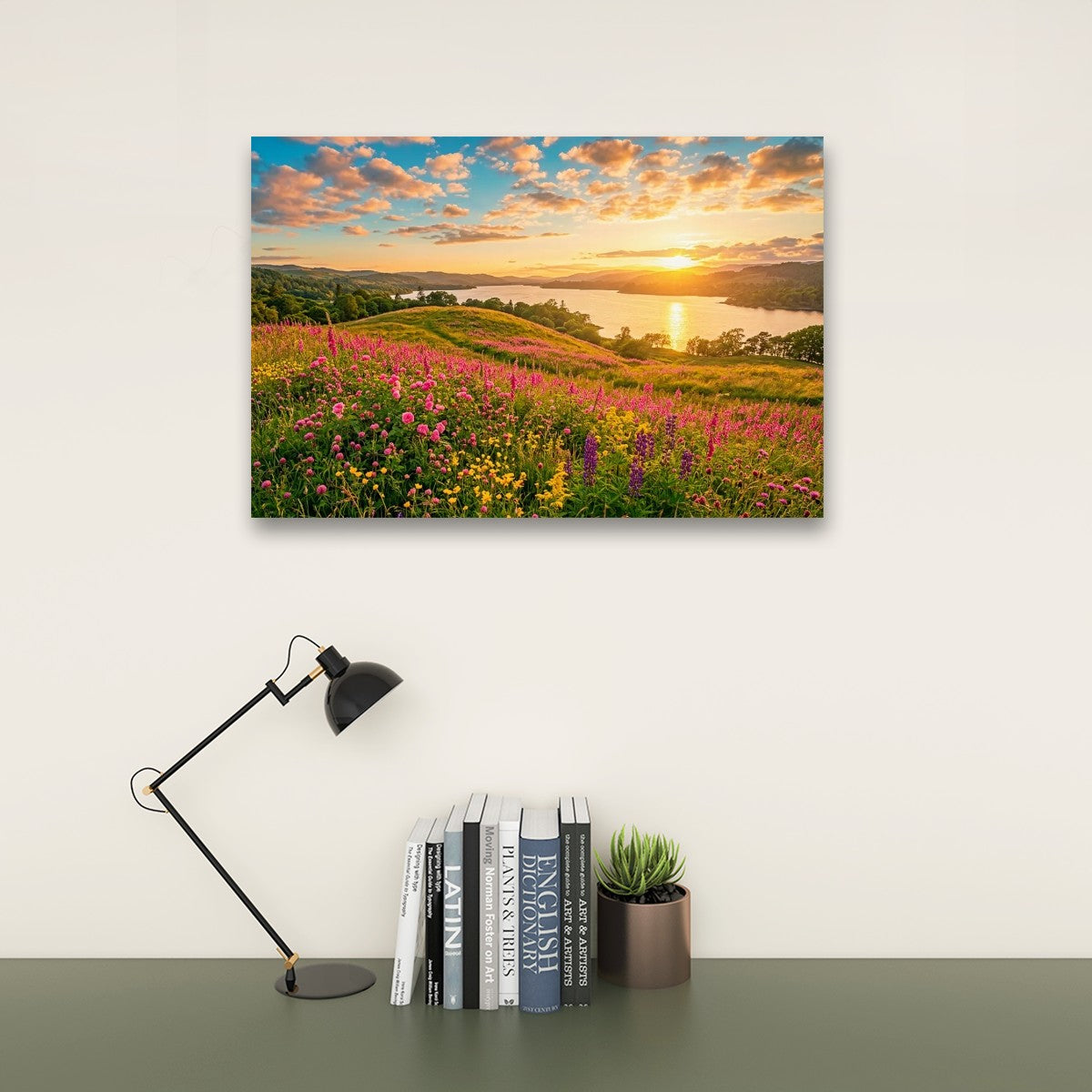 A rectangular wall art print showing a colorful wildflower meadow at sunset beside a lake, displayed on a white wall above a desk with a black lamp, books, and a small succulent.