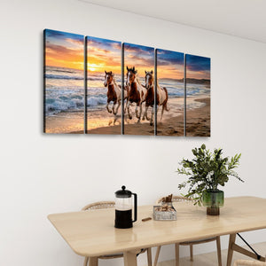 A five-panel canvas wall art set depicting three horses running along a beach at sunset, displayed on a white wall above a dining table with a plant and coffee maker.