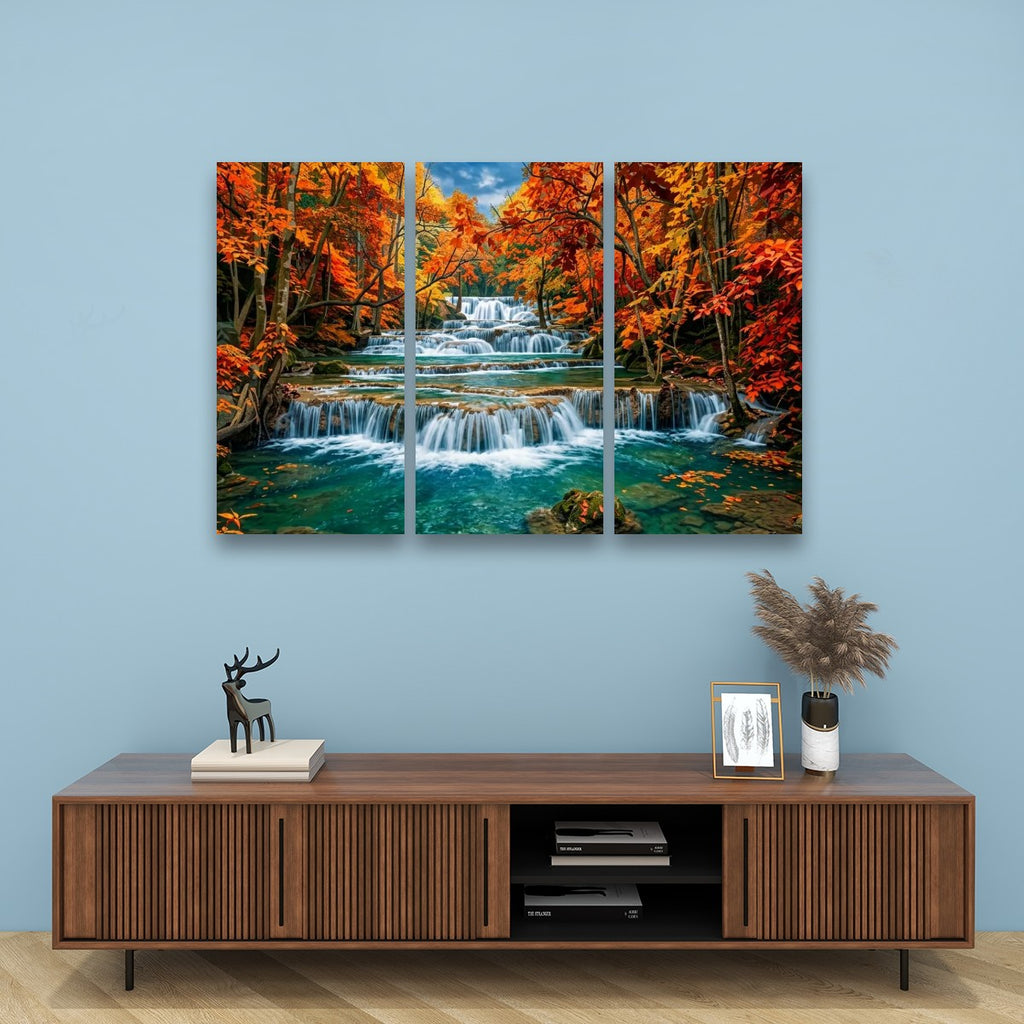 A three-panel canvas wall art print depicting a colorful autumn forest waterfall, displayed on a light blue wall above a wooden console table with decorative items.