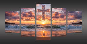 A five-panel canvas wall art set depicting a dramatic beach sunset with waves, glowing orange sky, and a large cross in the clouds, shown against a dark gray background.