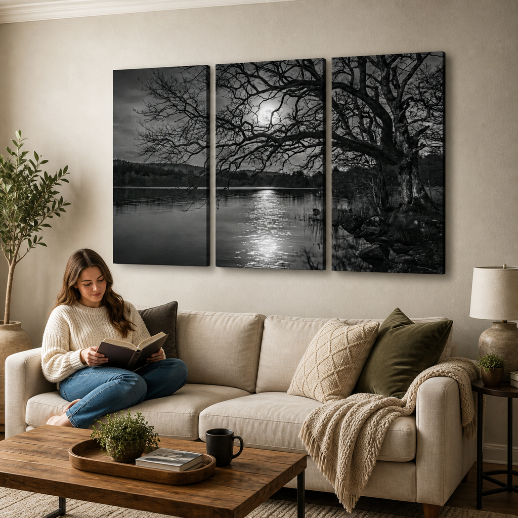 A woman sits on a beige sofa in a living room, reading a book. Above the couch is a three-panel black-and-white wall art print depicting a moonlit lake scene with bare trees and reflected light on the water.