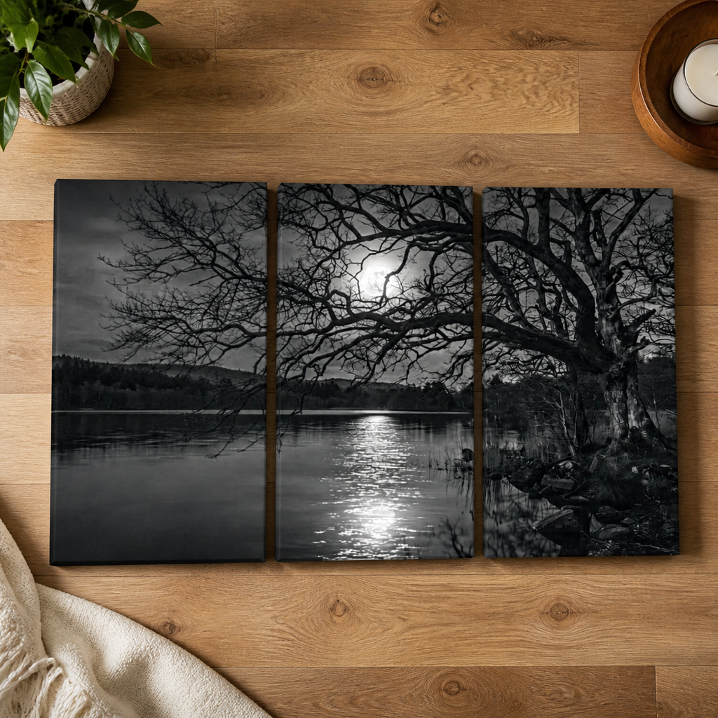 A three-panel black and white wall art print depicting a moonlit lake scene with bare tree branches, displayed on a wooden tabletop with a plant, candle, and blanket partially visible around it.