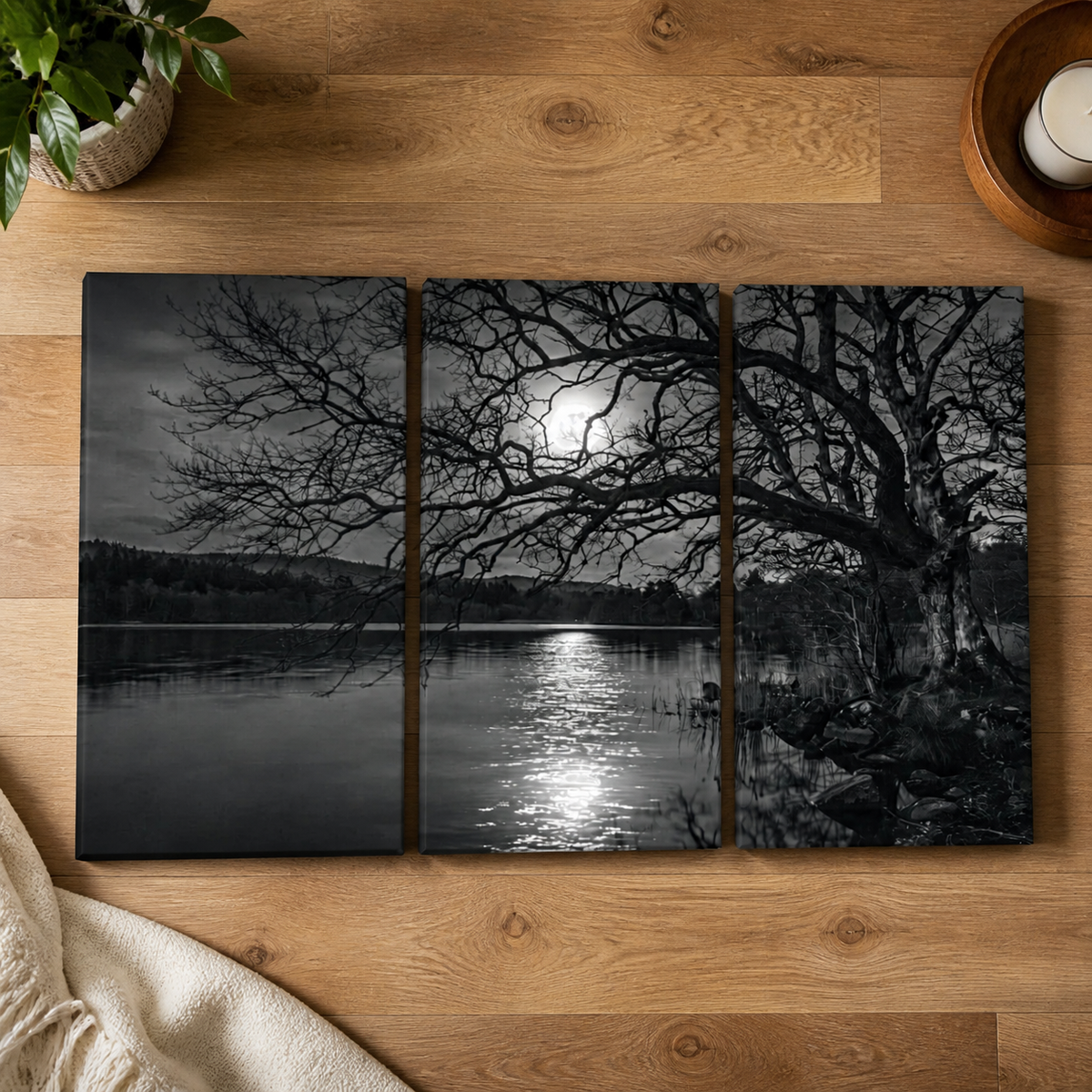 A three-panel black and white wall art print depicting a moonlit lake scene with bare tree branches, displayed on a wooden tabletop with a plant, candle, and blanket partially visible around it.