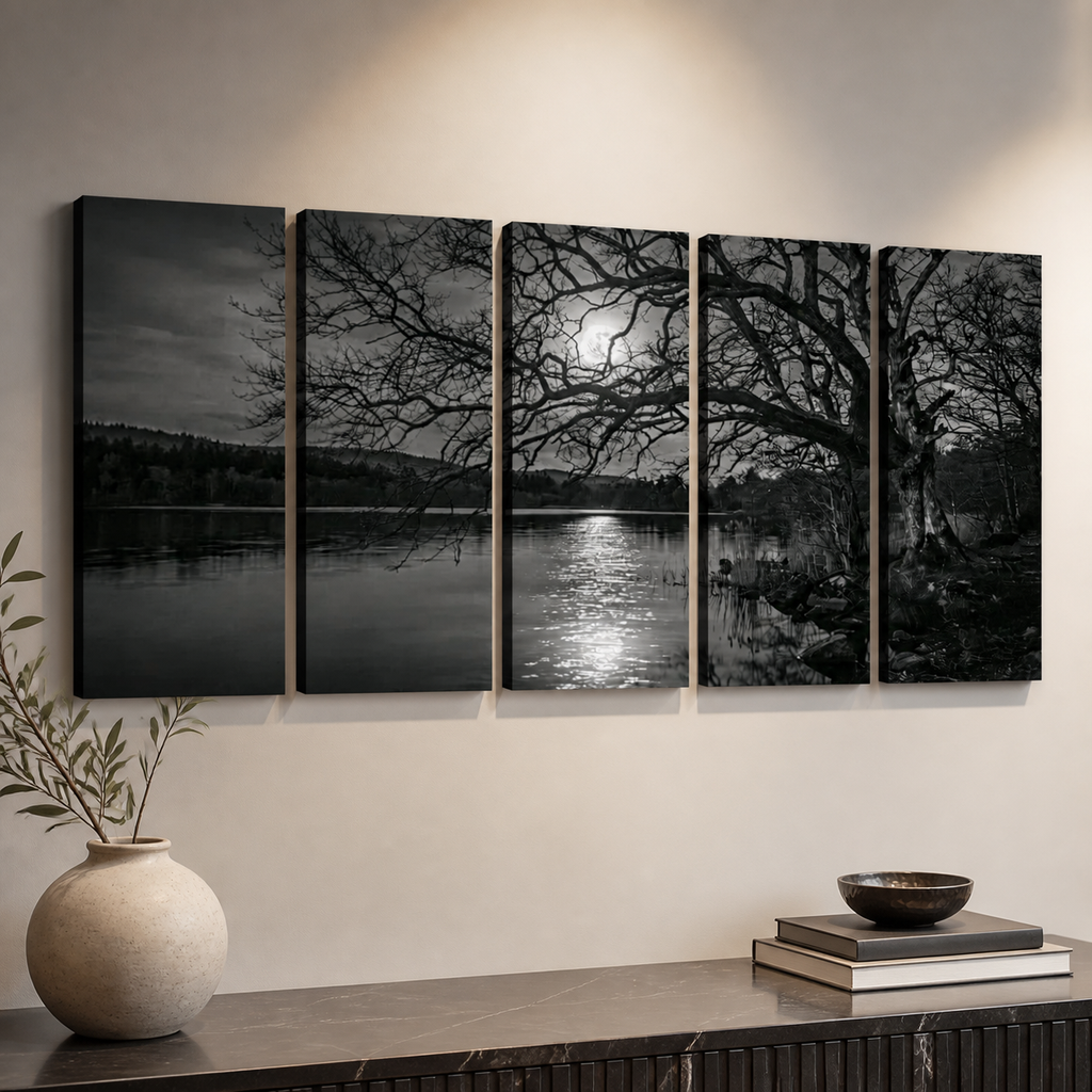 A five-panel black and white canvas wall art set depicting a moody lakeside scene with bare trees, a bright moon, and reflective water, displayed on a beige wall above a console table with a vase and books.