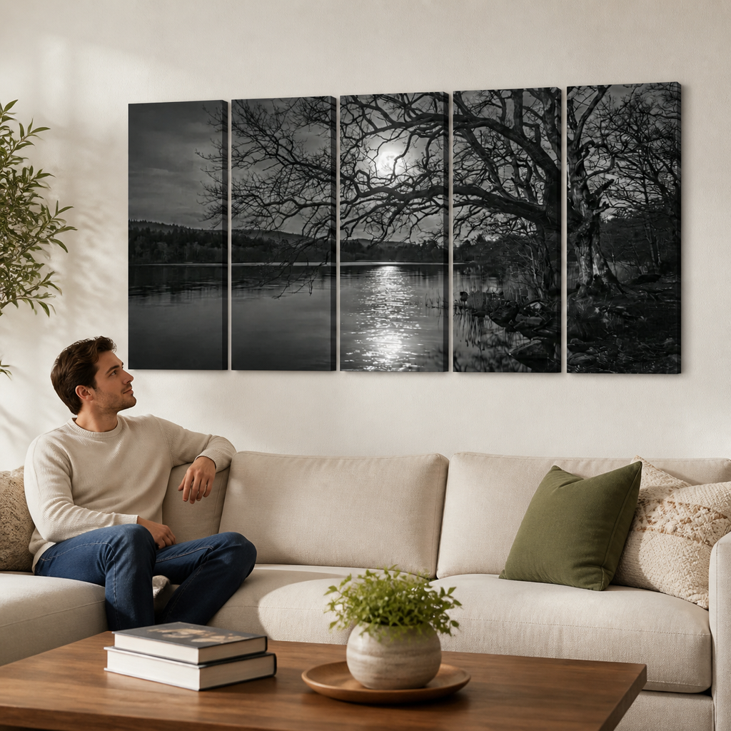 A man sits on a light beige sofa in a modern living room, looking at a large five-panel black and white canvas wall art set depicting a moonlit lake scene with bare trees.