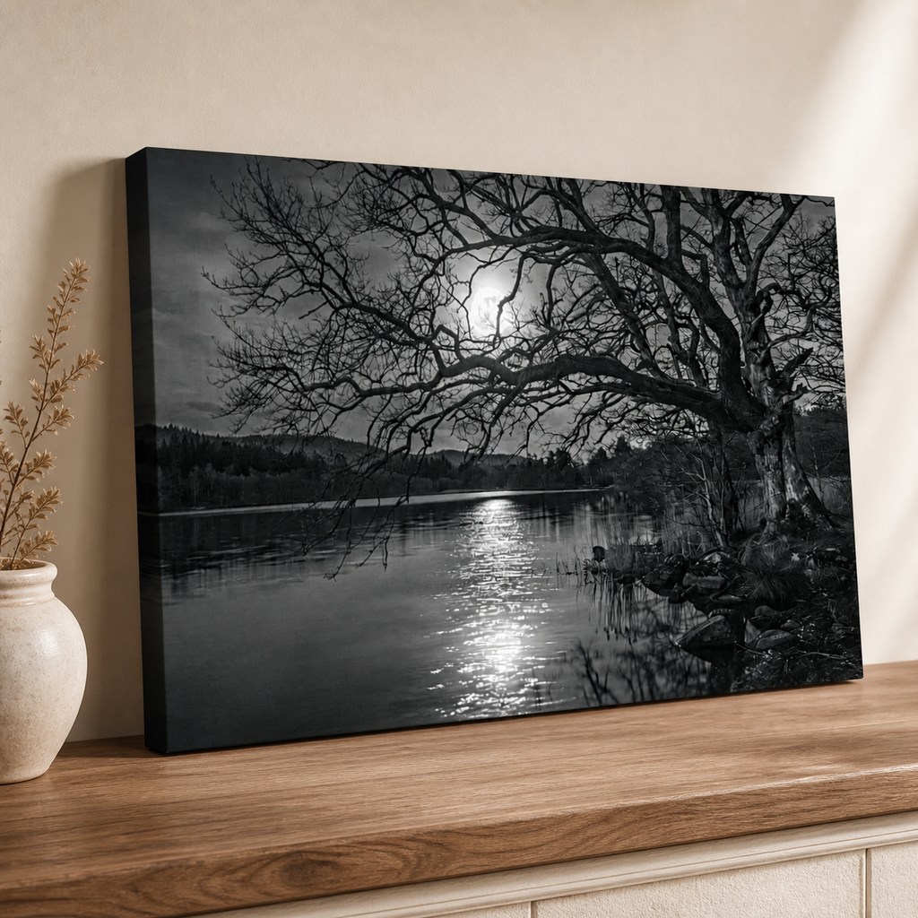 A black and white landscape canvas print of a moonlit lake scene with bare tree branches, displayed on a wooden mantel next to a white vase with dried stems.