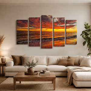 A 5-panel canvas wall art set depicting a dramatic orange and a red Indian Rocks,  FL sunset over the ocean, displayed above a modern beige sectional sofa in a styled living room.