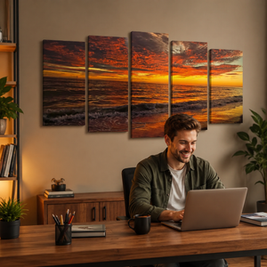 A man sits at a wooden desk using a laptop in a home office, with a five-panel canvas wall art set depicting a dramatic orange and yellow a Indian Rocks,  FL sunset over the ocean hanging on the beige wall behind him.