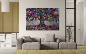 A three-panel canvas wall art set depicting a colorful tree with a dark brown trunk and vibrant purple, blue, pink, and green foliage, displayed on a white wall above a modern gray sofa in a bright living room.