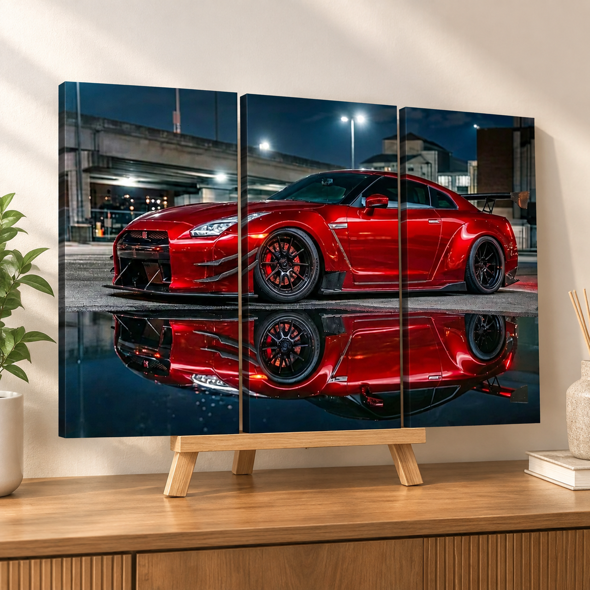 A three-panel canvas wall art print showing a red sports car reflected in water, displayed on a wooden stand on a console table in a styled room.