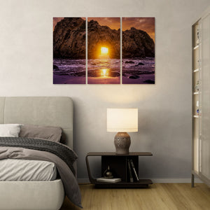 Triptych wall art of a sunset over water in a bedroom setting with a lamp and books on a nightstand.